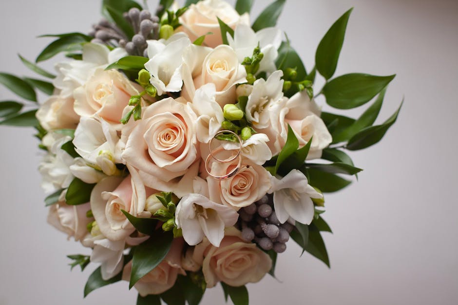 A close-up view of a floral bouquet featuring soft pink roses, white freesias, and small purple berries, complemented by lush green leaves and stems. The arrangement is beautifully handcrafted, with t